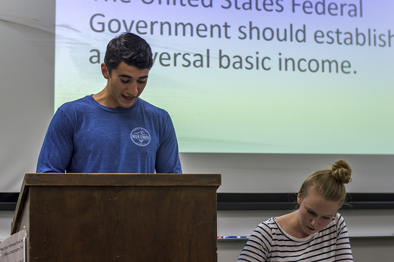 A student speaks at a podium in a debate round, with the motion on a universal basic income on the screen behind him